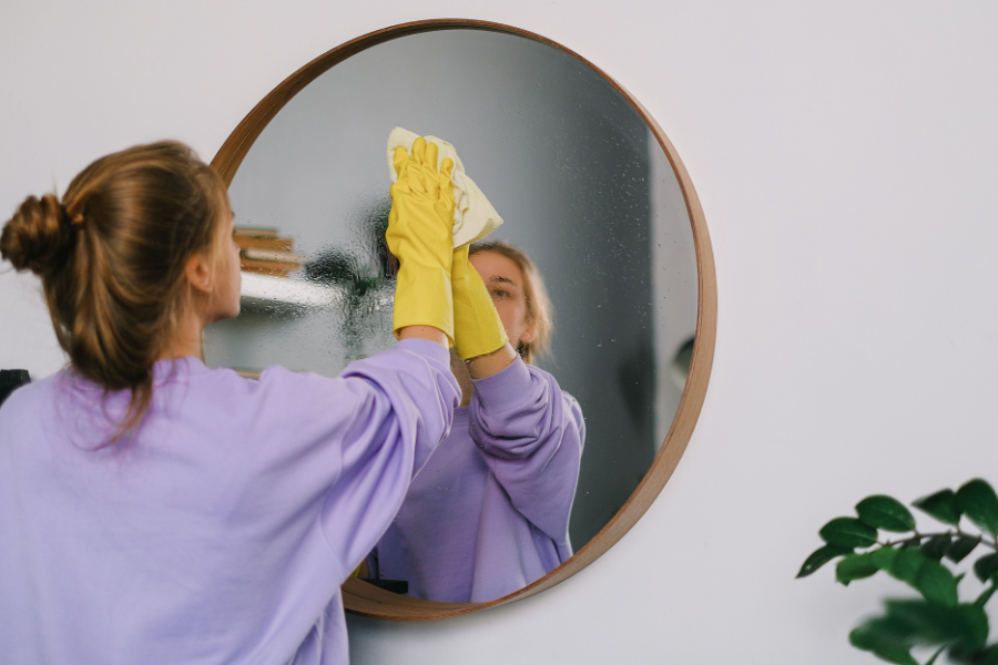 La imagen muestra a una mujer limpiando un espejo. Lleva guantes amarillos. Empleo hogar.